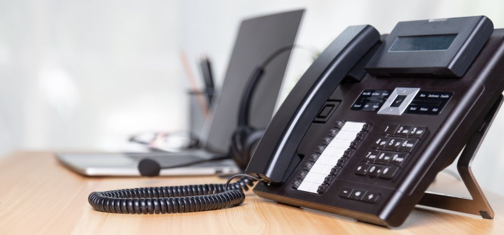 What to look for in a business phone system: A close-up of a black multi-line office desk phone on a wooden surface, with a blurred laptop, glasses, and stationery visible in the background.