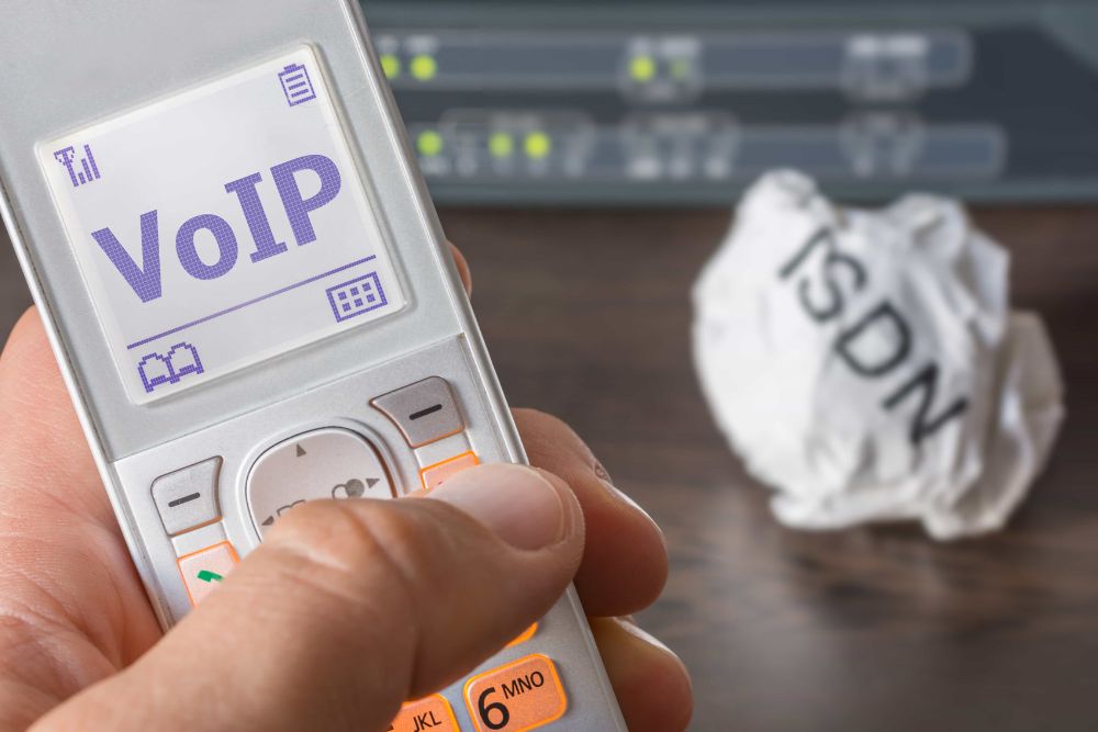 Business VoIP UK: A hand holding an old mobile phone displaying "VoIP" on its screen, with a crumpled piece of paper reading "ISDN" and network server equipment blurred in the background.