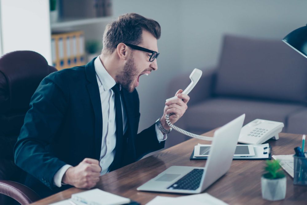 Is VoIP better than a landline: A frustrated man in glasses and a suit shouting into a corded telephone handset at his desk, with a laptop, a desk phone and office furniture around him.