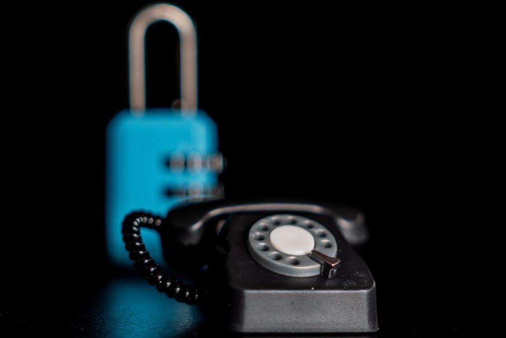 A black rotary dial telephone in the foreground with a blue combination padlock blurred behind it, both set against a dark background.