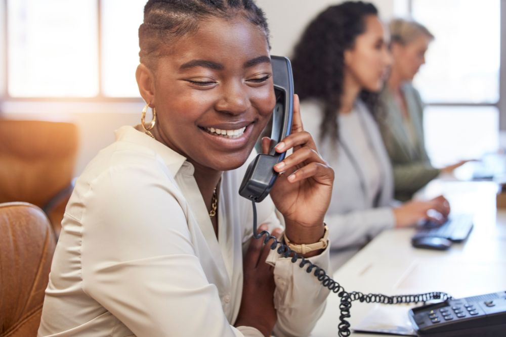 A smiling young woman in a white blouse holding a desk phone handset to her ear in an office, with two colleagues working in the blurred background.