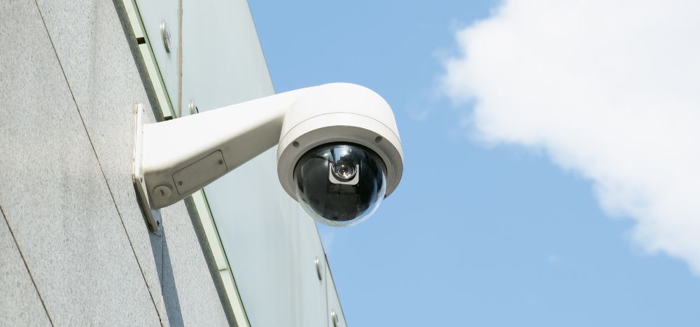 CCTV for business: White dome CCTV camera mounted on a bracket on a building wall against a blue sky.